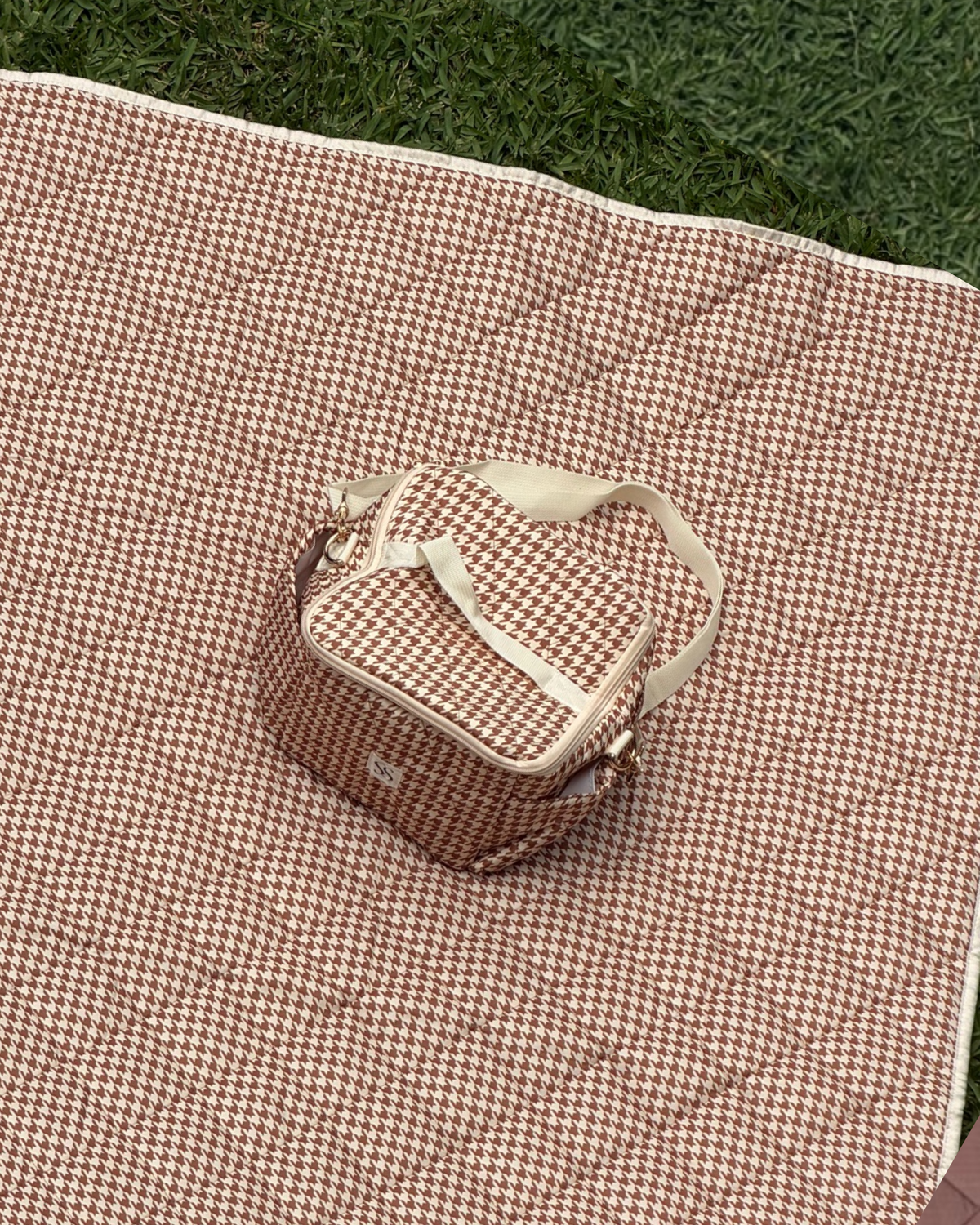 Biscuit cooler bag in terracotta and cream houndstooth with a cream shoulder strap, on the matching picnic blanket