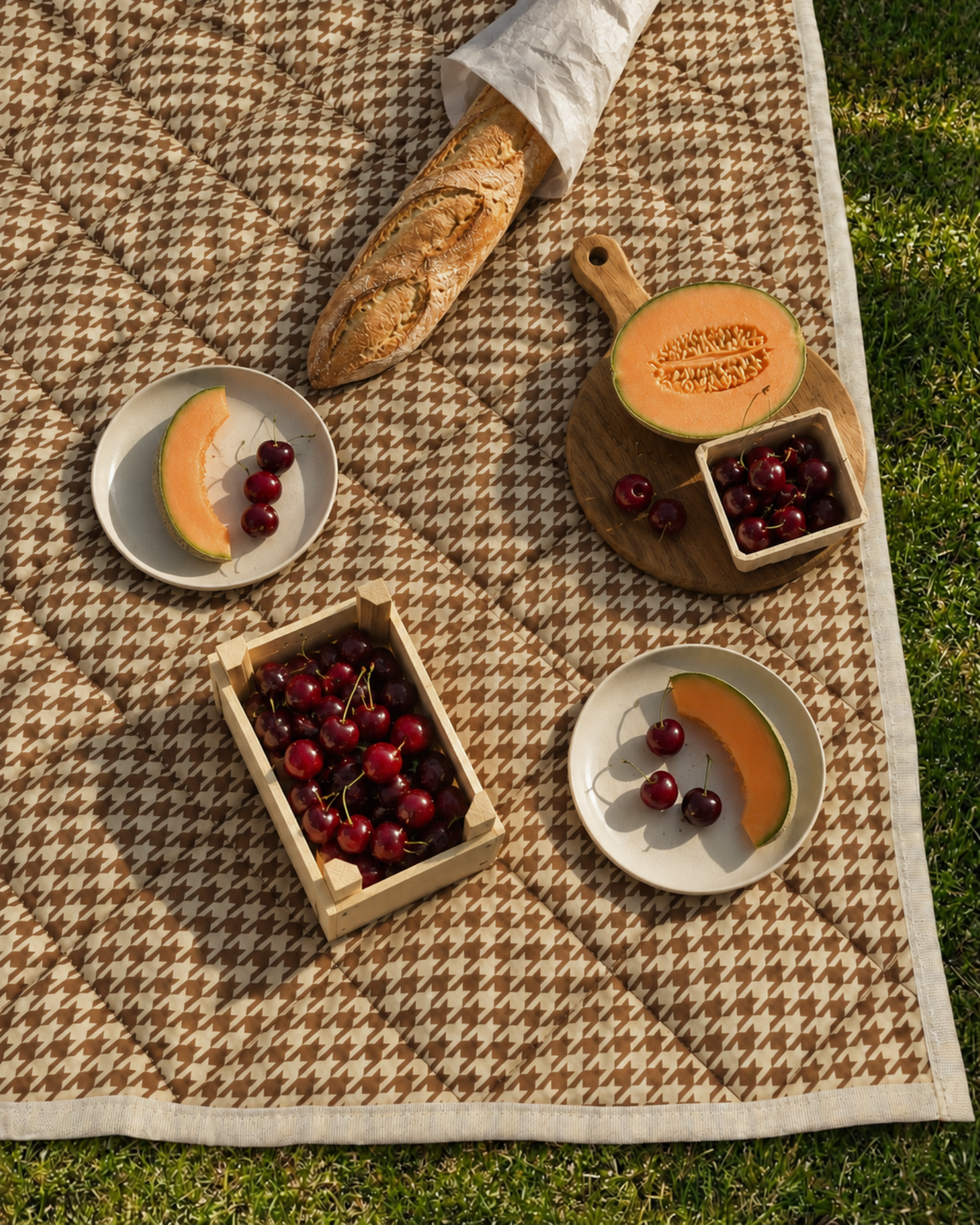 Biscuit picnic blanket in terracotta and cream houndstooth on the grass, set with cherries, rockmelon and a baguette