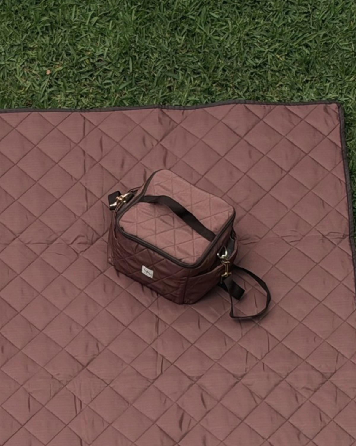 Chocolate gingham cooler bag with a zip top and brown shoulder strap, on the matching picnic blanket