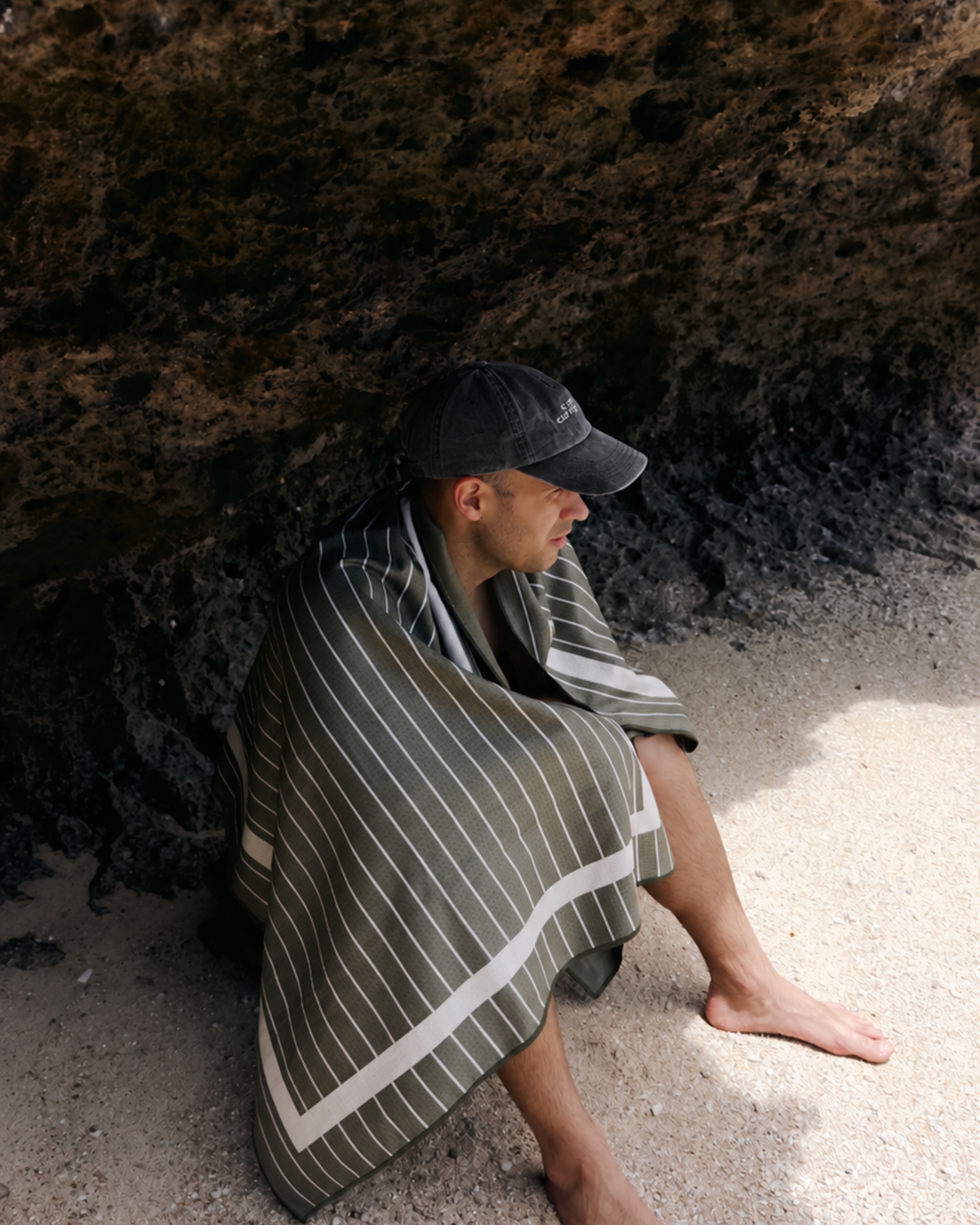 Man sitting on the sand in the shade of a rock wrapped in the Palm sand free beach towel, khaki with cream pinstripes
